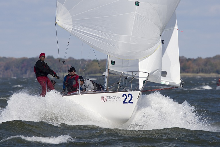 A white sailboat numbered 22 speeds through choppy water, creating splashes. Four people in red and dark clothing are onboard, sailing under a full white spinnaker with trees and sky in the background.