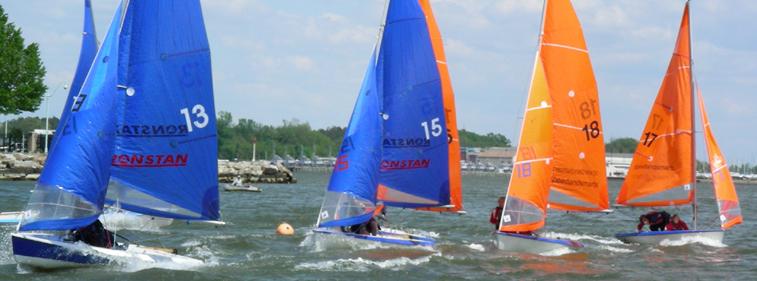 Four small sailboats, two with blue sails and two with orange sails, race on a windy lake. Sailors are leaning to balance the boats, and a tree-lined shoreline is visible in the background.