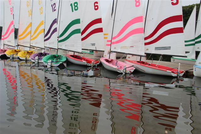 A row of colorful sailboats with striped sails and matching numbers is docked on calm water, creating a vibrant reflection of the boats and sails on the surface.