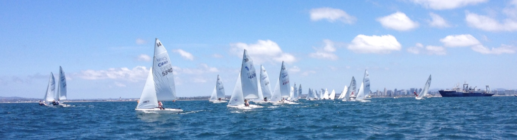 A group of small sailboats with white sails race on a blue ocean under a partly cloudy sky, with a city skyline and a large ship visible in the distant background.