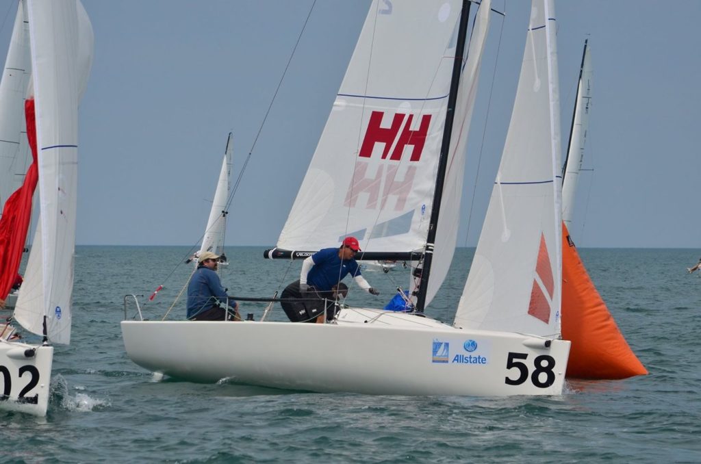 Three people sail a small white racing boat numbered 58 around an orange buoy on a calm sea, with other sailboats nearby under a clear sky.