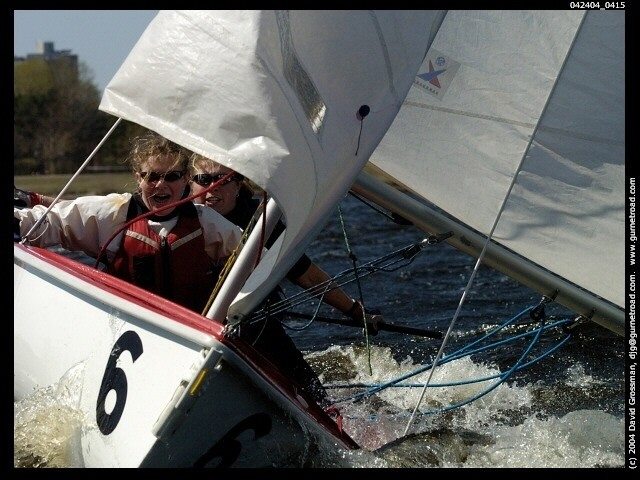 Two people wearing life jackets and sunglasses are sailing in a small sailboat with the number 6, leaning as the boat tilts sharply on the water, creating splashes and waves around them.