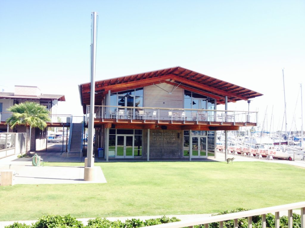 A modern two-story waterfront building with a wide terrace overlooks a marina with sailboats. The building is surrounded by green grass, palm trees, and clear blue sky.