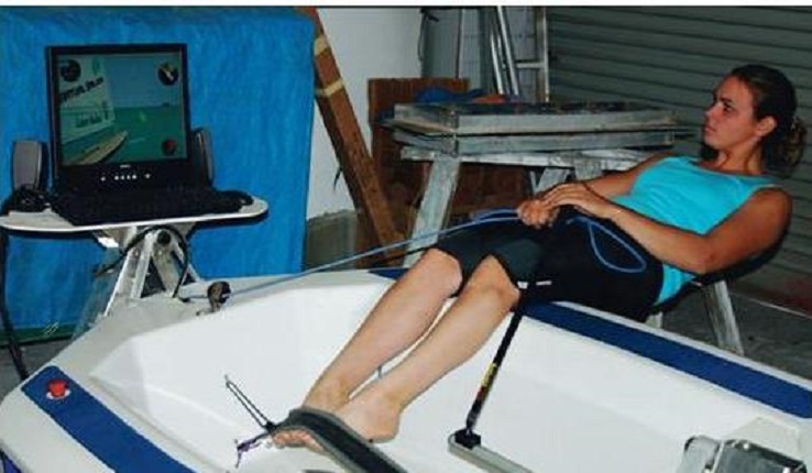 A woman sits in a small boat indoors, holding ropes as she looks at a computer screen in front of her, simulating rowing or sailing. The setup is in a garage or workshop with tools and tarps in the background.