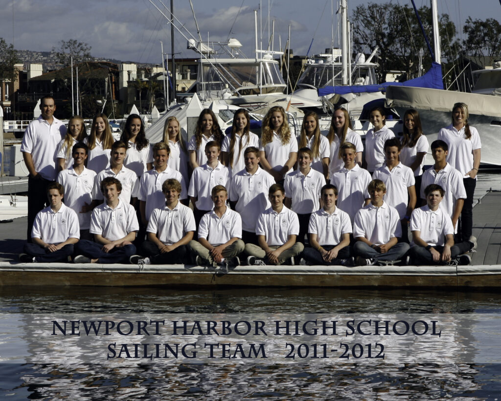 A group of students in white shirts pose on a dock in front of boats for the Newport Harbor High School Sailing Team photo, 2011–2012. The team is seated and standing in rows, with a marina and water in the background.