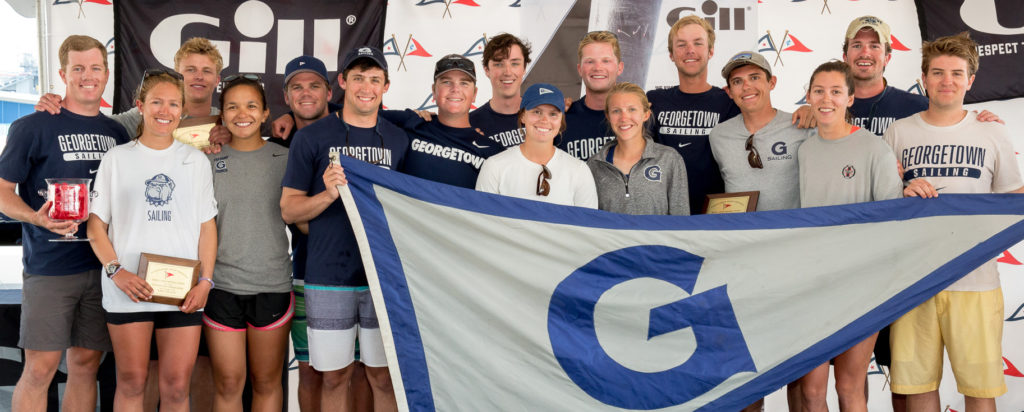 A group of young adults in Georgetown Sailing shirts and shorts pose together holding plaques and a large Georgetown flag, smiling in front of a branded backdrop at an awards event.