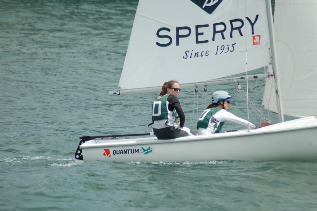 Two people wearing life vests and numbered bibs are sailing a small white boat on the water. The sail displays the word 'SPERRY' and the side of the boat reads 'QUANTUM Sails.