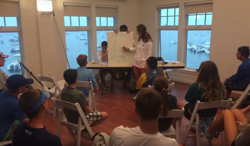 A group of young people sit in a semicircle in a room with large windows overlooking water. Two stand at the front, one pointing at a diagram on a whiteboard as others watch and listen attentively.