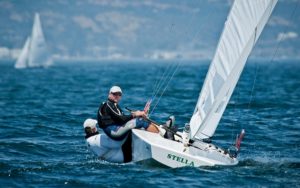 Two people sailing a small white boat named "Stella" on blue water, with one person leaning out over the edge. Both wear wetsuits and sunglasses. Other sailboats and a blurred coastline are visible in the background.