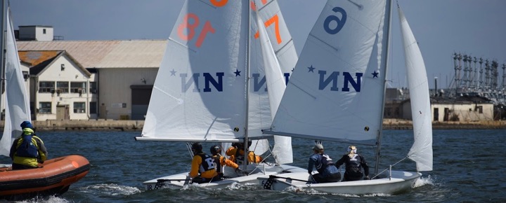 Two small sailboats with teams of sailors maneuver closely in a race near a large industrial building, while a person in a safety boat watches from the water. The sails display numbers and 'N' logos.