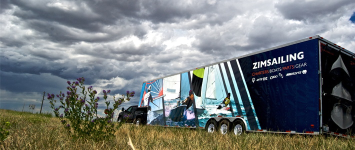 A truck with a colorful “ZimSailing” trailer featuring images of people sailing is parked in a grassy field under a dramatic, cloudy sky. Purple wildflowers grow in the foreground.