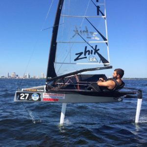 A man sails alone in a small hydrofoil sailboat with the number 27, gliding above the water on foils. City buildings and a clear blue sky are visible in the background.