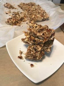 A stack of homemade granola bars with nuts and seeds on a white plate, with more granola bars on parchment paper in the background.