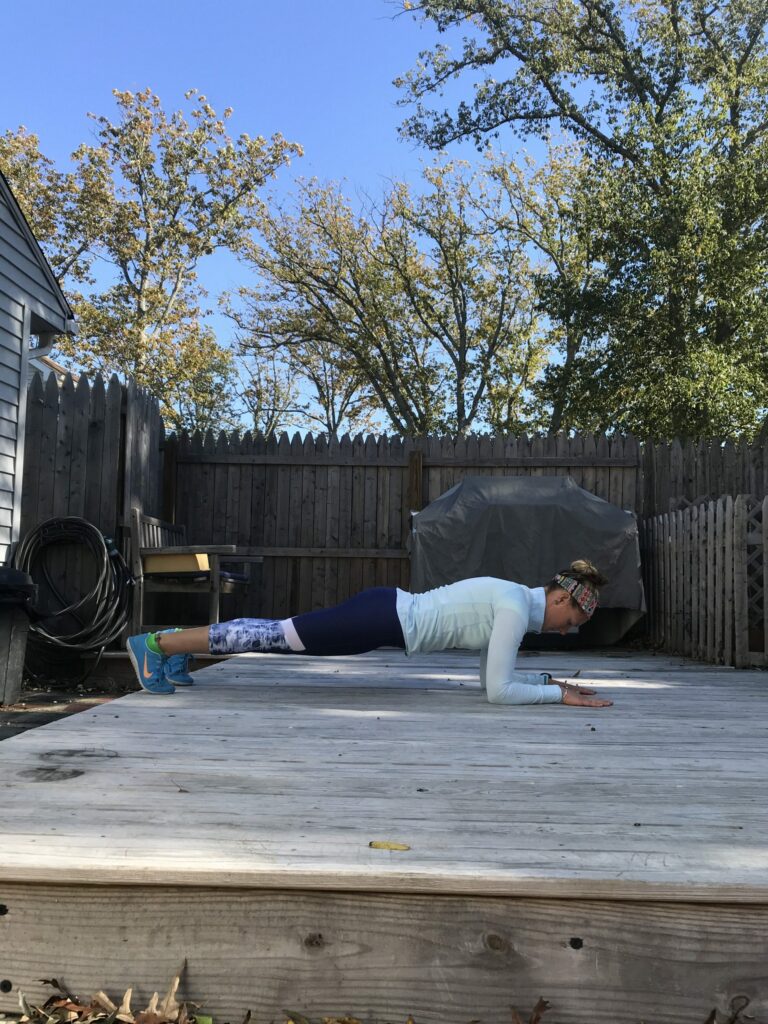 A person in athletic clothing performs a forearm plank on a wooden deck outside, with a fence, trees, and grill visible in the background on a sunny day.