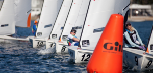 Several sailors compete in a race, maneuvering small sailboats around an orange buoy marked "Gil" on open water, with their sails angled closely together. The sailors wear life vests and sunglasses.
