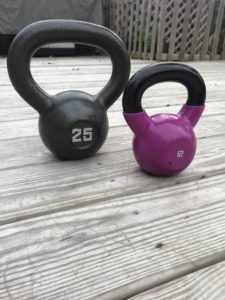 A 25-pound gray kettlebell and a 12-pound purple kettlebell are placed side by side on a wooden deck outdoors.