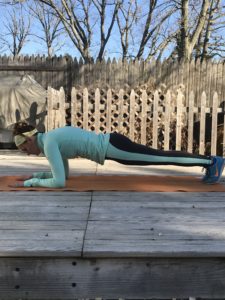 A person wearing a light blue long-sleeve shirt, headband, and leggings holds a forearm plank position on an orange mat outdoors on a wooden deck, with a wooden fence and bare trees in the background.