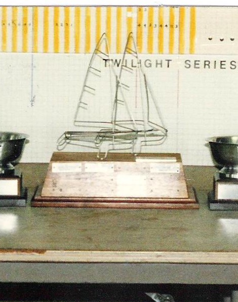 A metal wire sailboat trophy on a wooden base is displayed on a table between two silver cups. Behind them, a board reads 'TWILIGHT SERIES' with a grid and yellow stripes at the top.