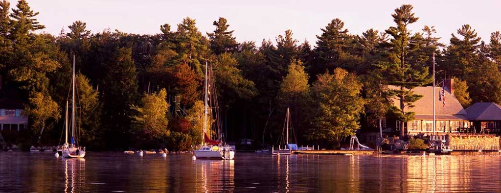 Sailboats are anchored near a wooden dock beside a building surrounded by tall trees, all reflected in the calm lake water during sunset. The scene is peaceful with warm golden light.