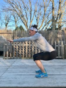 A woman wearing a gray long-sleeve shirt, black shorts, and blue sneakers performs a squat exercise outdoors on a wooden deck with a wooden fence and leafless trees in the background.