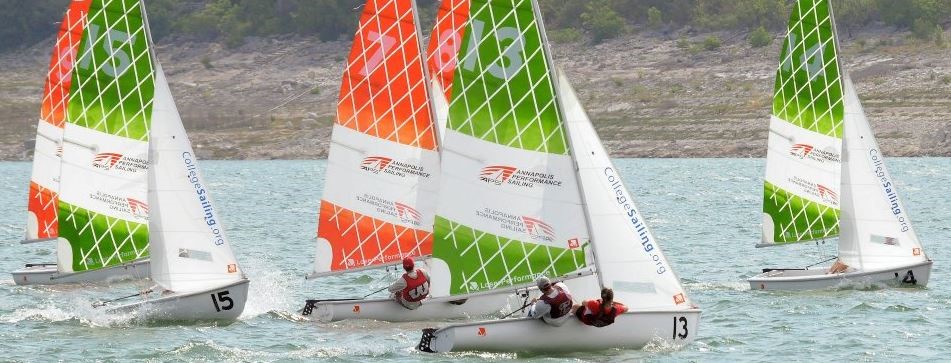 Several small sailboats with colorful green and orange patterned sails race on a lake, with teams of sailors in each boat and a rocky shoreline with trees in the background.