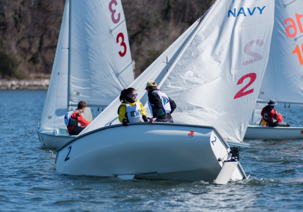 Two sailboats with visible 'NAVY' and number '2' on their sails compete on a sunny day. Four sailors are aboard, wearing life jackets and gear, navigating close to each other on calm water. Trees are in the background.