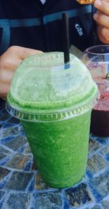 A green smoothie in a clear plastic cup with a lid and straw sits on a mosaic table. In the background, a person in a dark jacket holds food, and another drink with a purple color is partially visible.