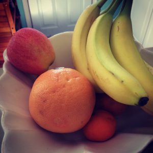A white bowl filled with bananas, an apple, a grapefruit, and two small oranges sits on a table with a door in the background.