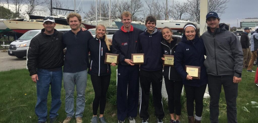 A group of seven people stands together outdoors, smiling and holding award plaques. There are sailboats and cars in the background, and everyone is dressed in jackets and casual attire. It appears to be a team event.