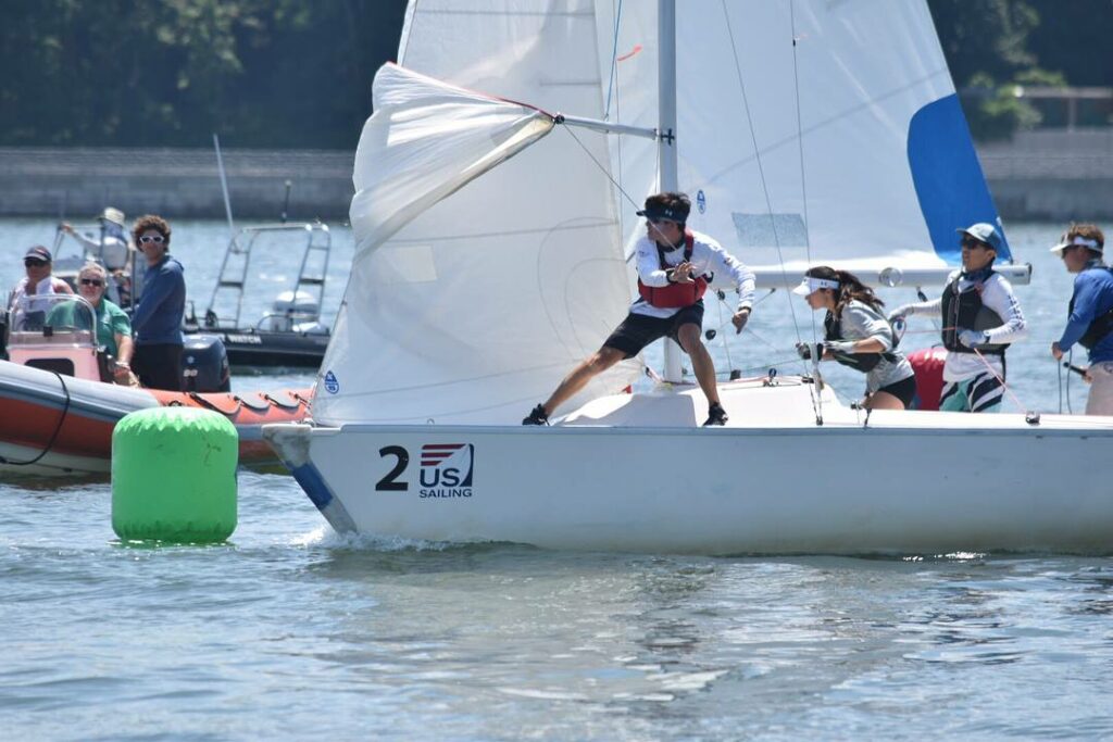 A sailboat with three crew members racing past a green buoy, marked '2 US Sailing,' as people watch from nearby boats on a sunny day.