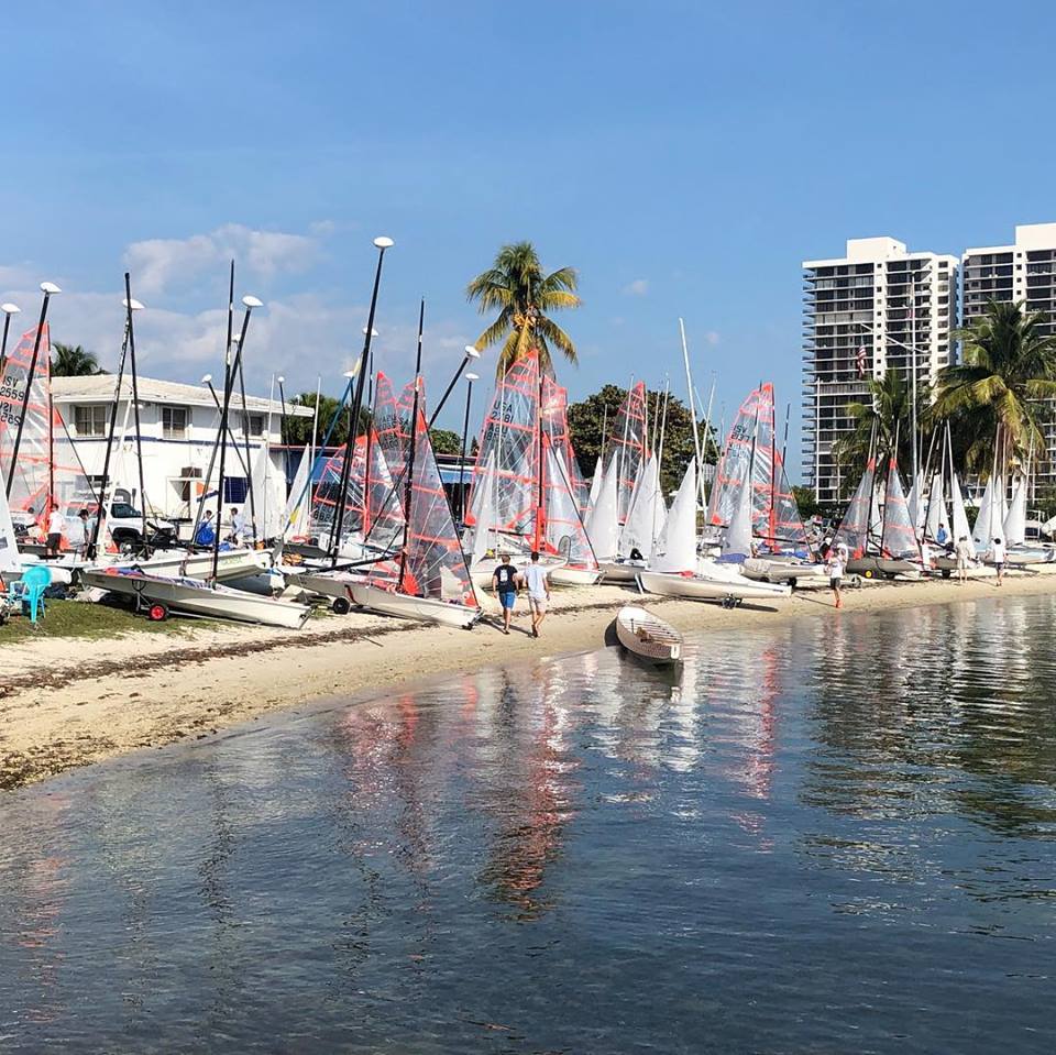 A sandy shoreline with several sailboats lined up, people preparing boats, palm trees, and tall modern buildings in the background under a clear blue sky.