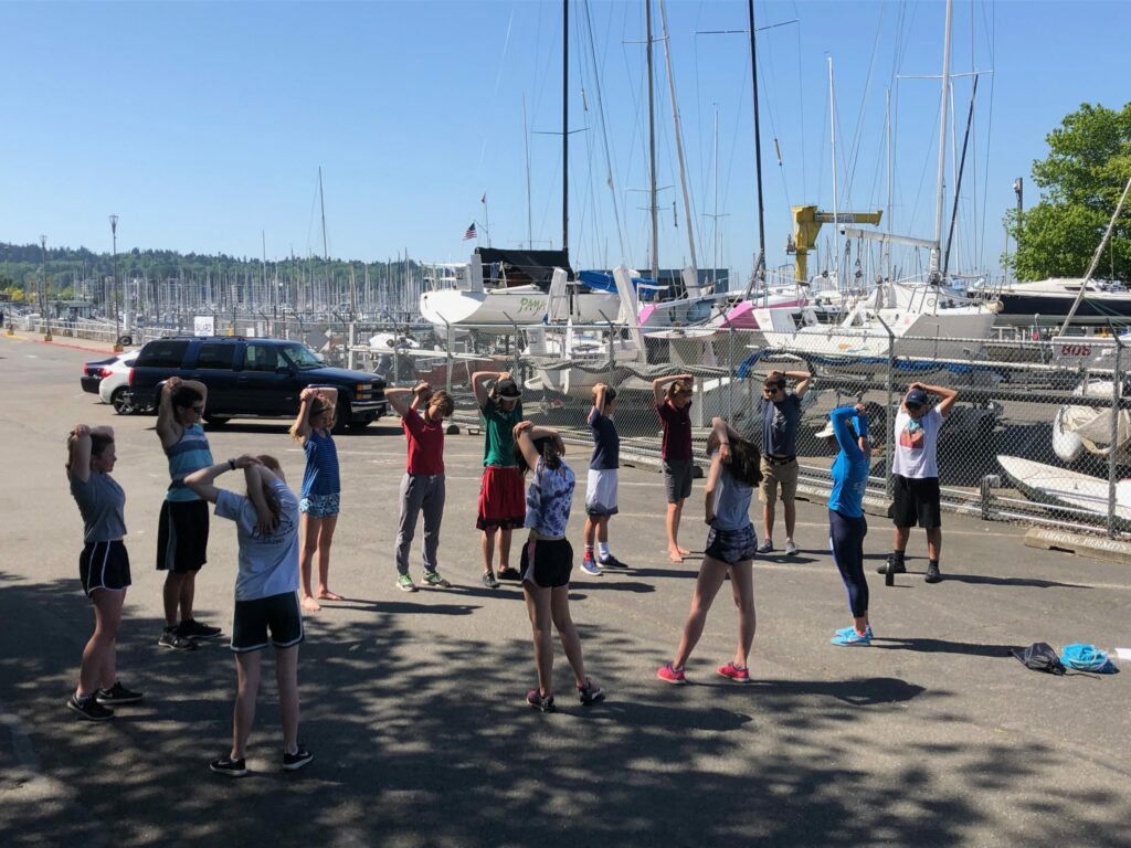 A group of teenagers stands in a circle on a sunny marina lot, stretching with hands behind their heads. Sailboats and cars are parked in the background near a chain-link fence.