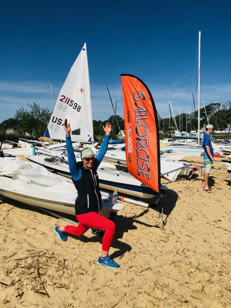 A person in bright athletic clothing kneels on sandy ground with arms raised, smiling near a 'SAILORSJC' flag and sailboats, under a clear blue sky. Another person stands nearby among the boats.