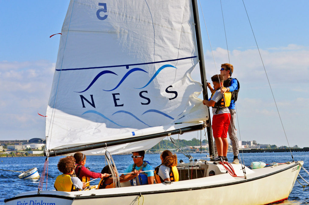 Six people wearing life jackets sail a small boat on a sunny day. One person adjusts the sail labeled 'NESS' while others sit or steer. The water is calm, and buildings and trees are visible in the background.