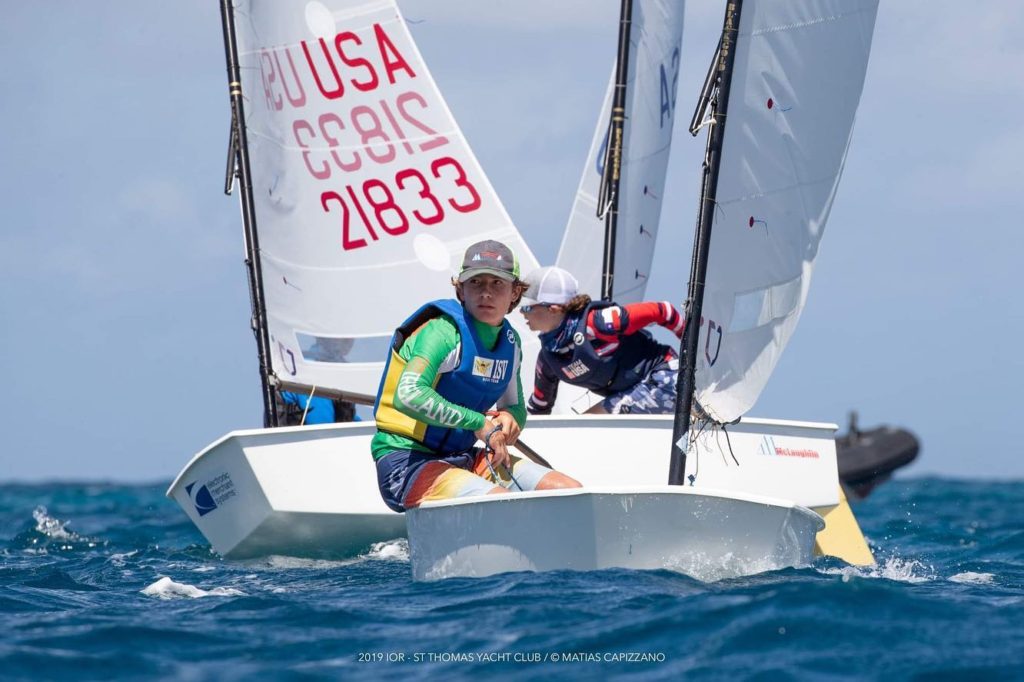 Two young sailors compete in small dinghy sailboats on blue water, with one wearing a green and yellow vest and the other in a dark top. The sails display numbers and 'USA.' The sky is clear and sunny.