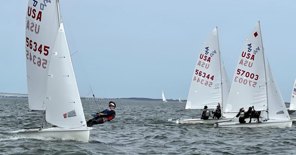 Three small sailboats with USA sail numbers race on choppy water. One sailor leans far out over the edge of their boat, while the others steer. The sky is overcast and a distant bridge is visible.