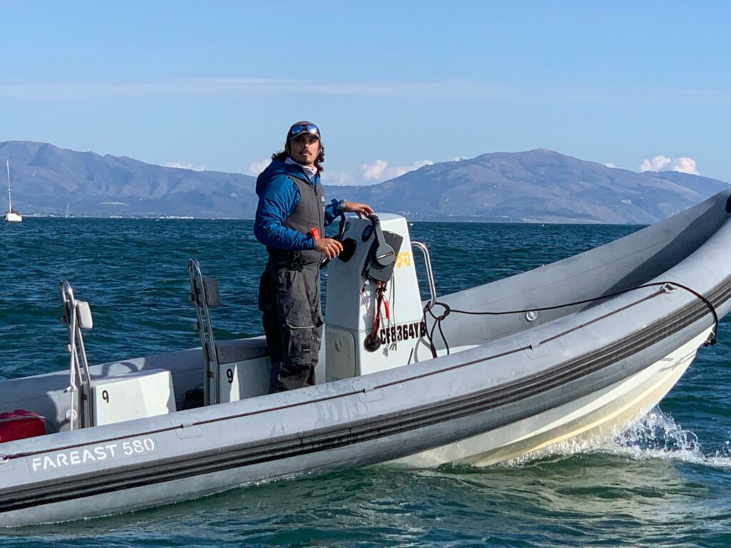 A person wearing sunglasses and a jacket stands at the control of a small inflatable boat on a body of water, with mountains and a sailboat visible in the background under a clear sky.