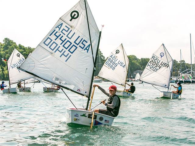 Several young sailors compete in small white sailboats on a calm lake, each boat displaying unique sail numbers. Trees and boats are visible in the background under a bright sky.