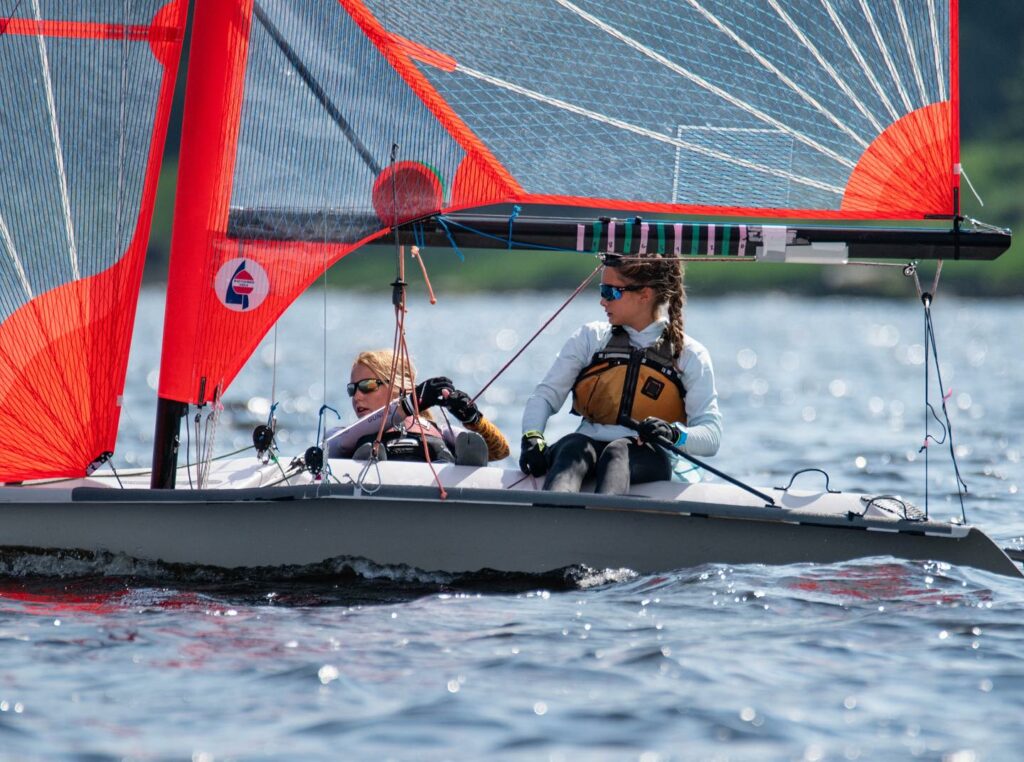Two people wearing life jackets sail a small boat with bright red sails on a calm body of water. One person steers while the other adjusts a rope, both focused on sailing. Trees are visible in the blurry background.