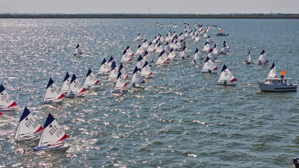 A large group of small sailboats with white sails race across a sunlit body of water, closely aligned in rows, with a motorboat on the right displaying an orange flag.
