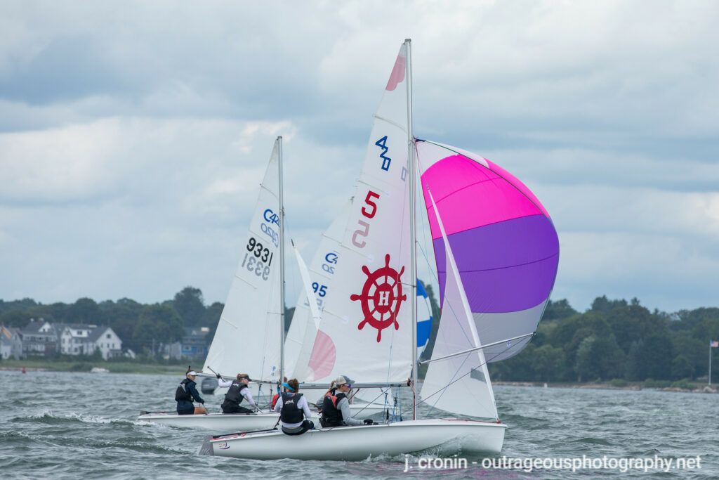 Two sailboats race on the water, with one boat displaying a pink and purple spinnaker and a red ship wheel symbol on its sail. A shoreline with houses and trees is visible in the background under a cloudy sky.