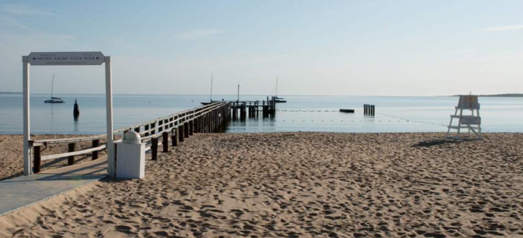 A sandy beach leads to a wooden pier extending over calm water, with a few boats anchored nearby. A lifeguard chair sits on the right, and the sky is clear and blue.