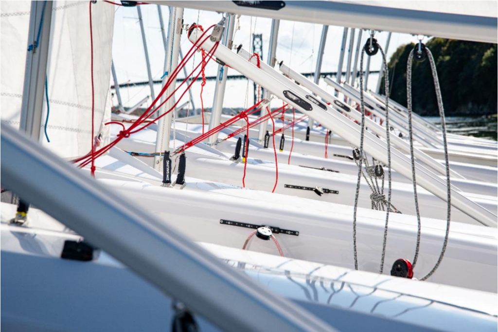 A close-up view of several white sailboats lined up side by side, showing their angled masts, ropes, and rigging, with a blurred natural background and water visible.
