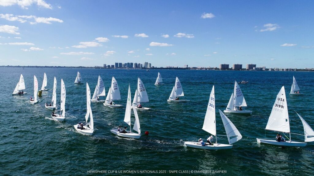 A fleet of small sailboats with white sails races across a blue body of water under a sunny sky, with a city skyline visible in the distance.