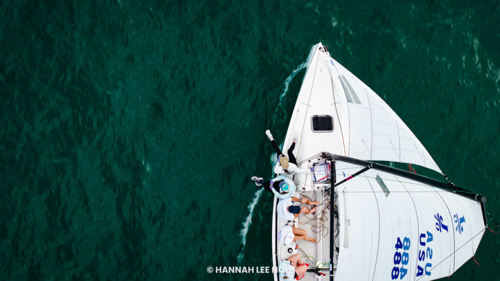 Aerial view of a white sailboat with several people on board sailing on deep green water. The boat's sail has “USA” and “488” written on it.