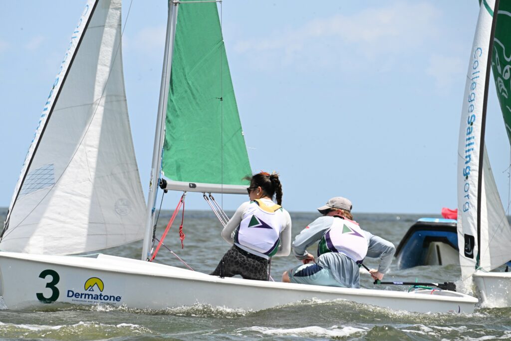 Two sailors wearing life vests race a small sailboat marked with the number 3 on the water. The boat has green and white sails, and there is another boat visible in the background under a blue sky.