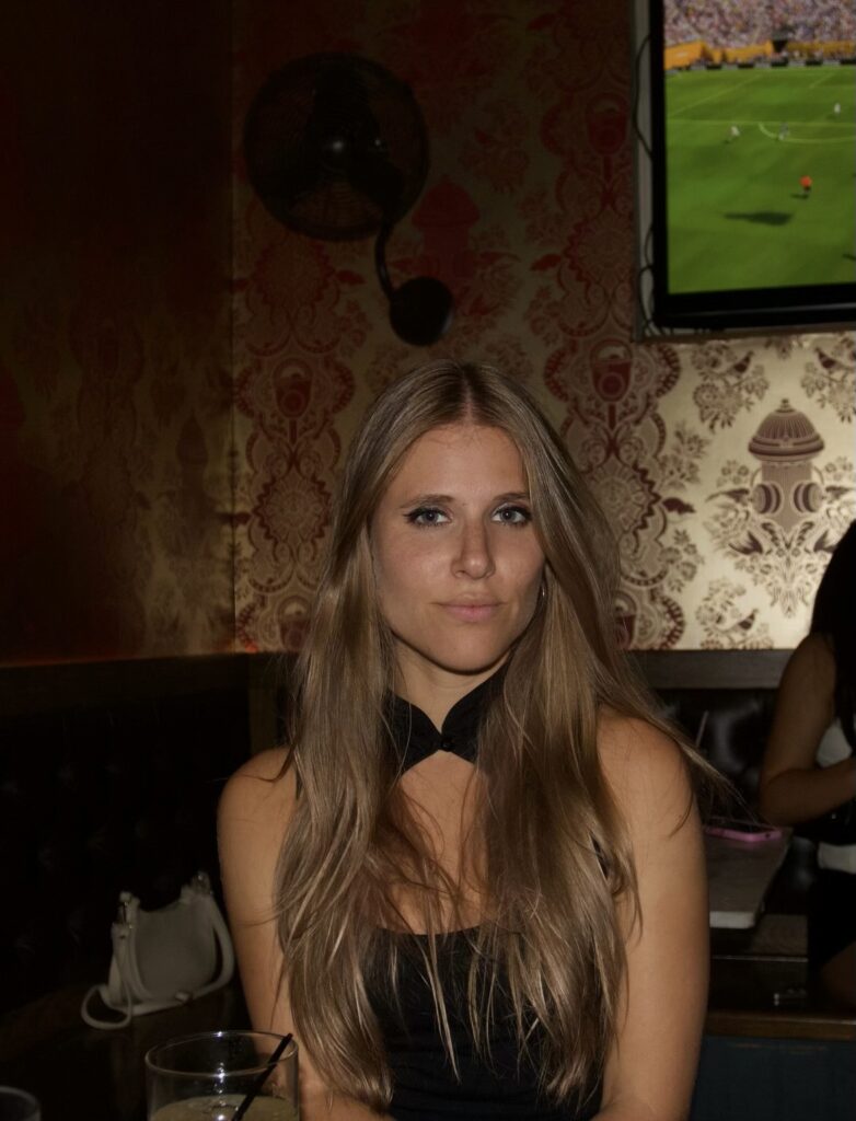 A woman with long, straight blonde hair sits at a table in a dimly lit bar. She is wearing a sleeveless black top. A sports game is playing on a TV in the background.