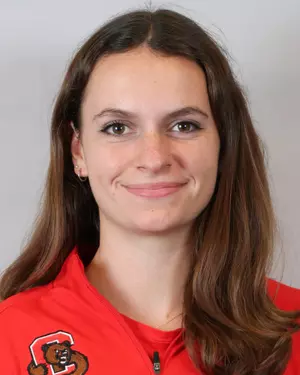 A young woman with long brown hair wearing a red jacket, smiling at the camera in front of a plain light background.