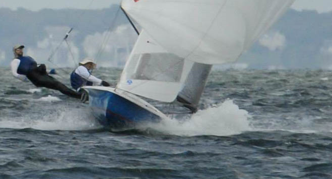 Two people sail a small boat in choppy water, leaning back to balance as the boat tilts and splashes through waves; a shoreline with houses is visible in the blurred background.