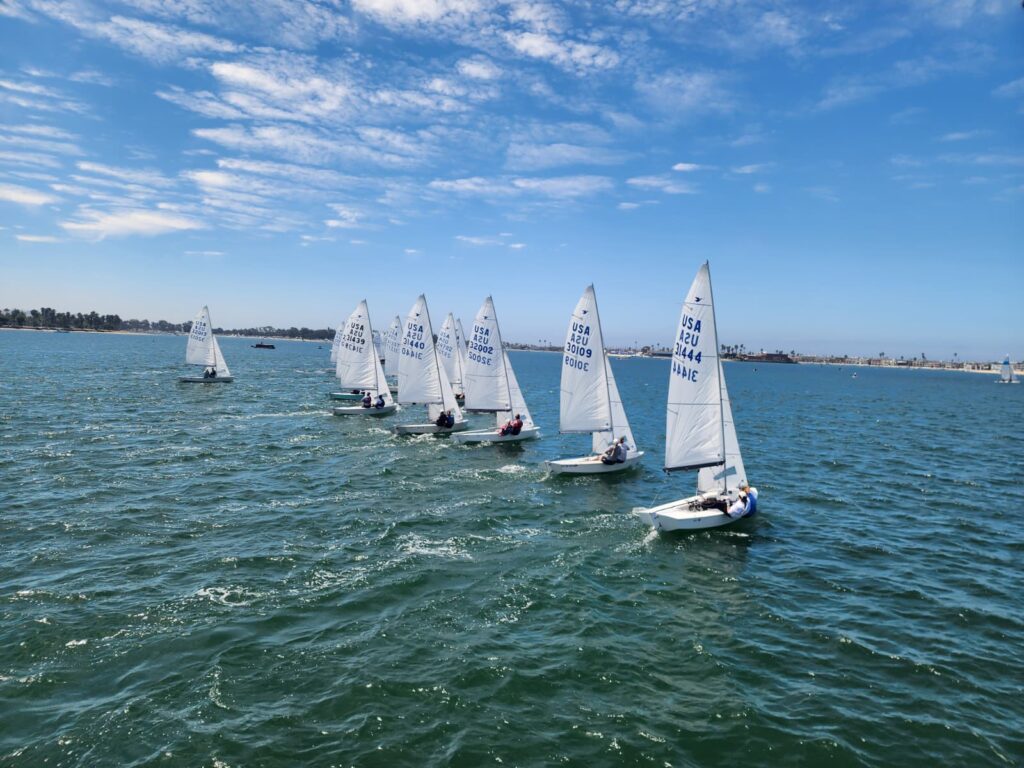 Seven small sailboats with white sails race across blue water under a partly cloudy sky. The boats are spaced in a line, sailing toward the right. The shoreline and some trees are visible in the background.